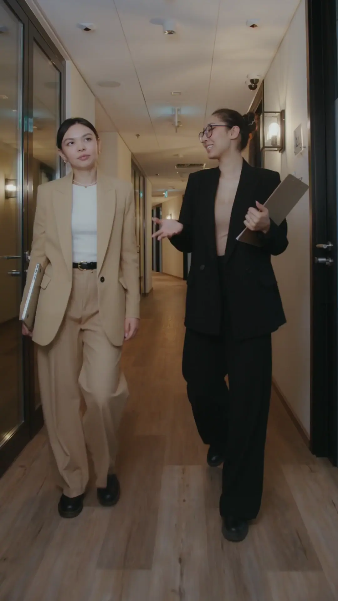 Two professional women in business suits walk down a brightly lit office corridor with wooden flooring.