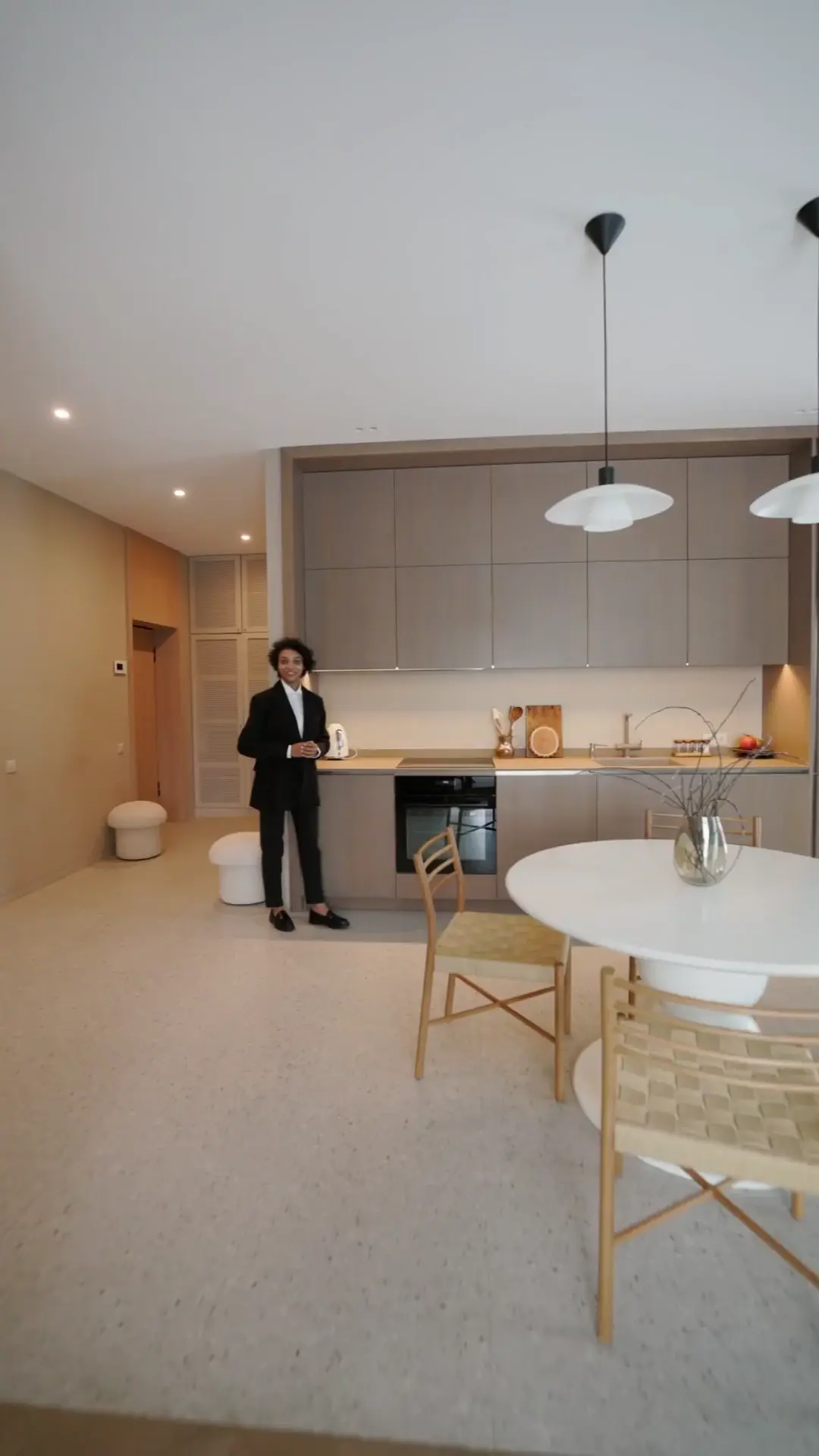 clipzy - image -01 A woman in a black pantsuit stands in a modern, minimalist kitchen with beige cabinets and a built-in oven.