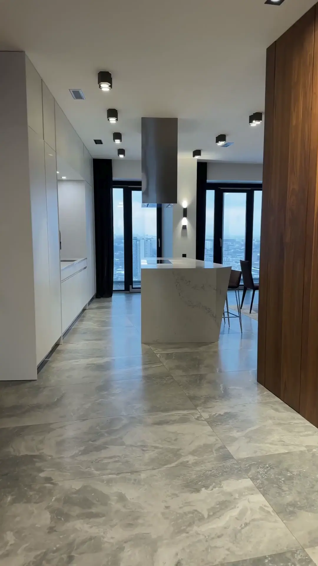 clipzy - image 03 A modern penthouse kitchen features glossy gray marble floor tiles and a central white marble waterfall island with a stainless steel range hood.